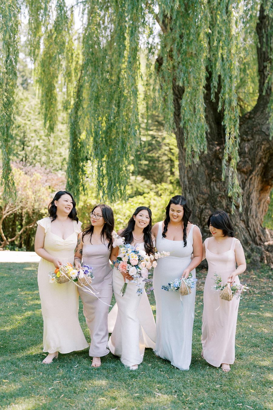 A group of bridesmaids in pastel dresses walking on a lush green lawn beneath a large willow tree, each holding a colorful bouquet, creating a joyful and serene wedding atmosphere.