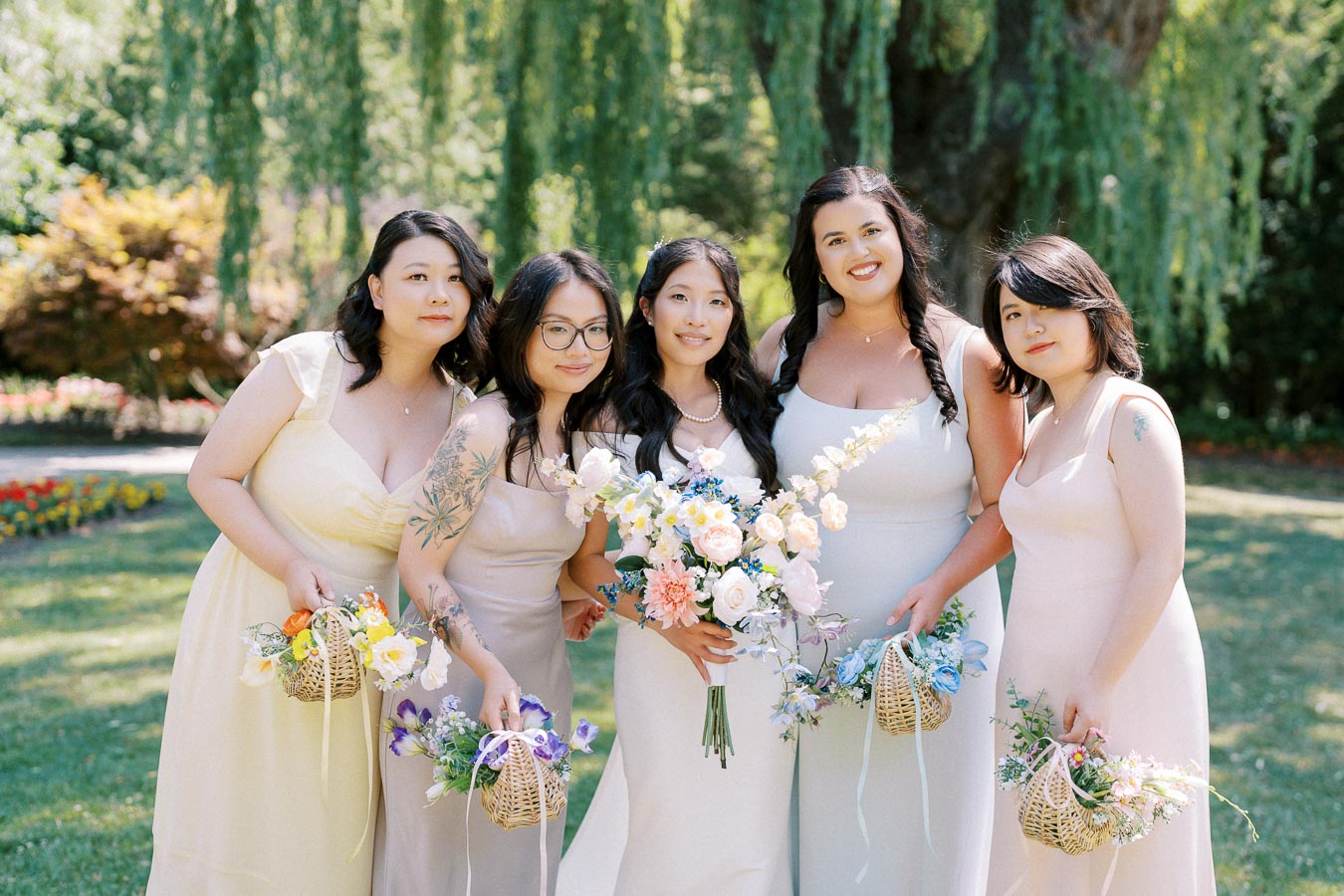 Five bridesmaids in pastel dresses holding floral baskets, posing outdoors in a garden setting with lush greenery and a weeping willow tree.