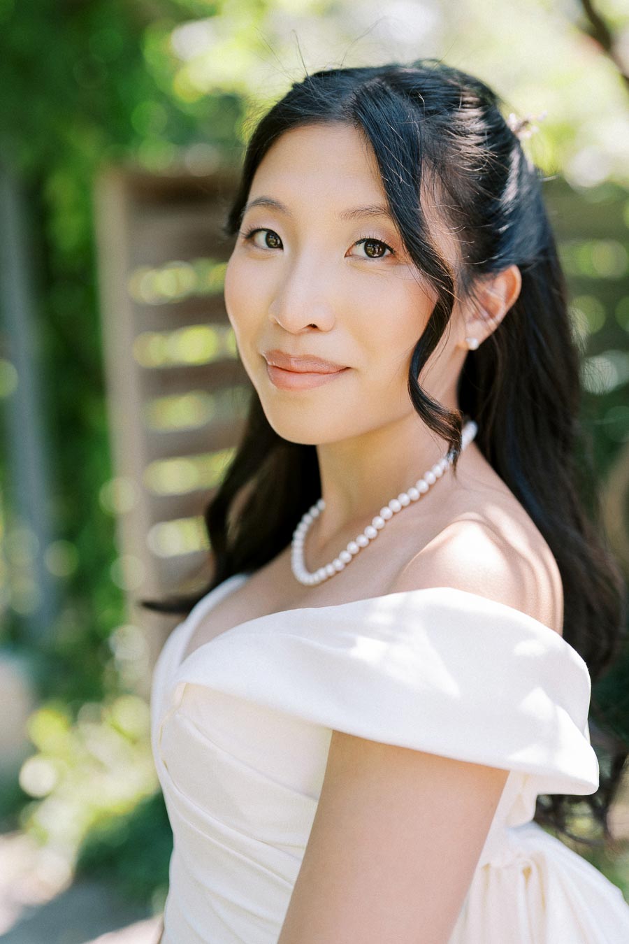 Bride wearing an elegant white off-shoulder wedding dress with pearl necklace, smiling softly with a lush green outdoor background.