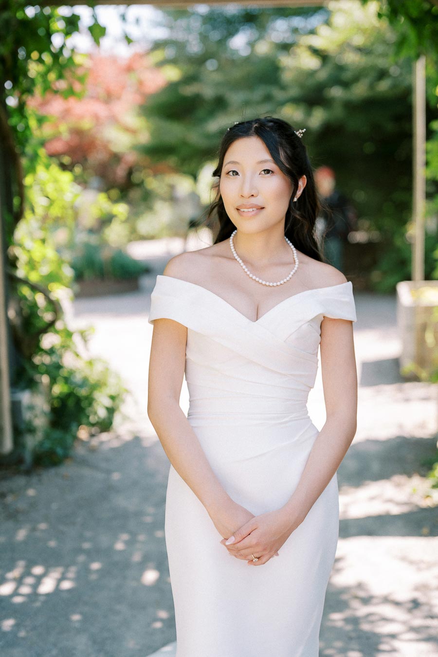 A bride in an elegant off-shoulder wedding dress stands outdoors, wearing a pearl necklace with a serene expression. The background features greenery and soft sunlight, creating a romantic and peaceful setting.