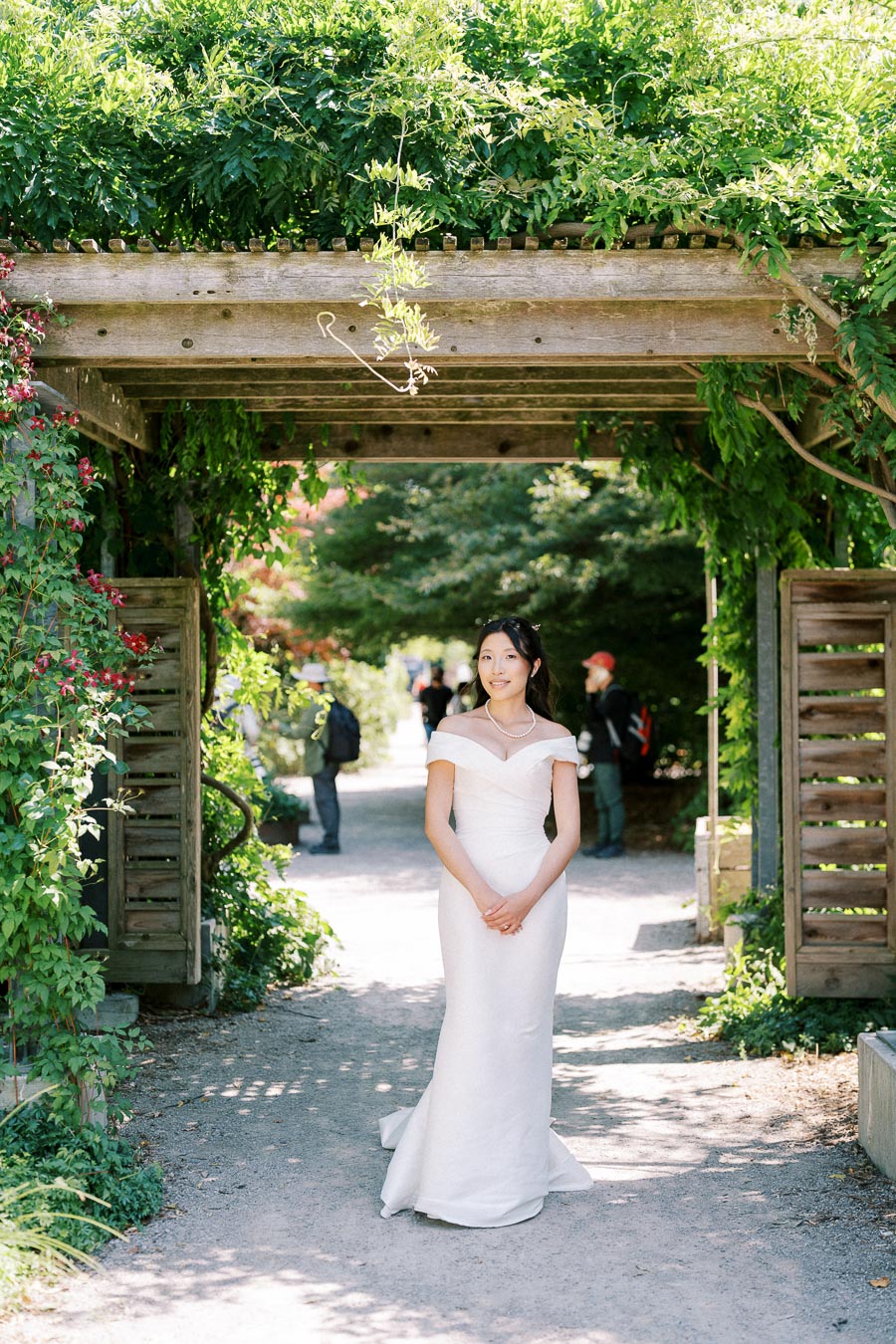 Bride in elegant white dress standing under a wooden pergola adorned with lush green foliage in a garden setting.