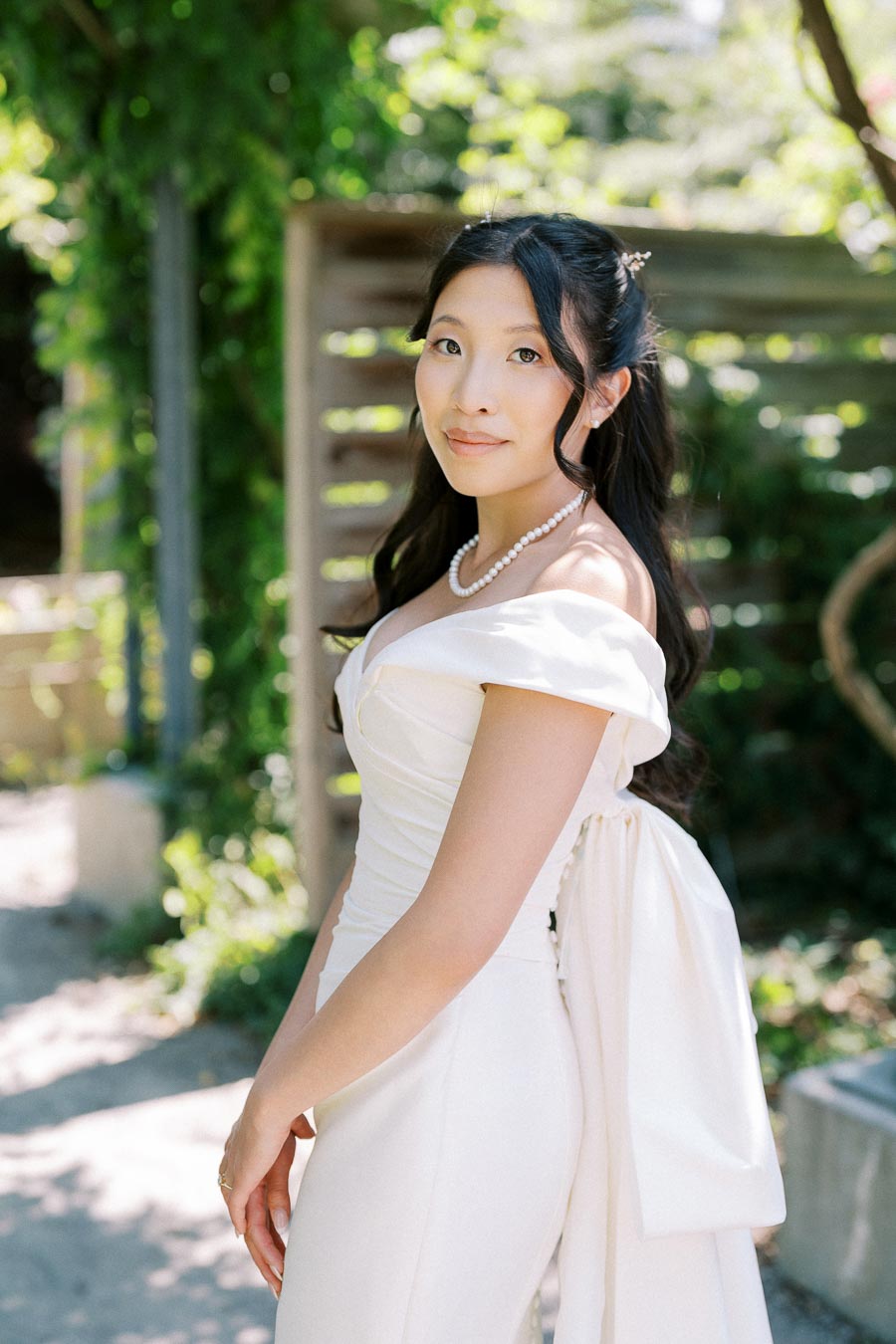A bride in an elegant white dress and pearl necklace stands outdoors, surrounded by lush greenery, on a sunny day.