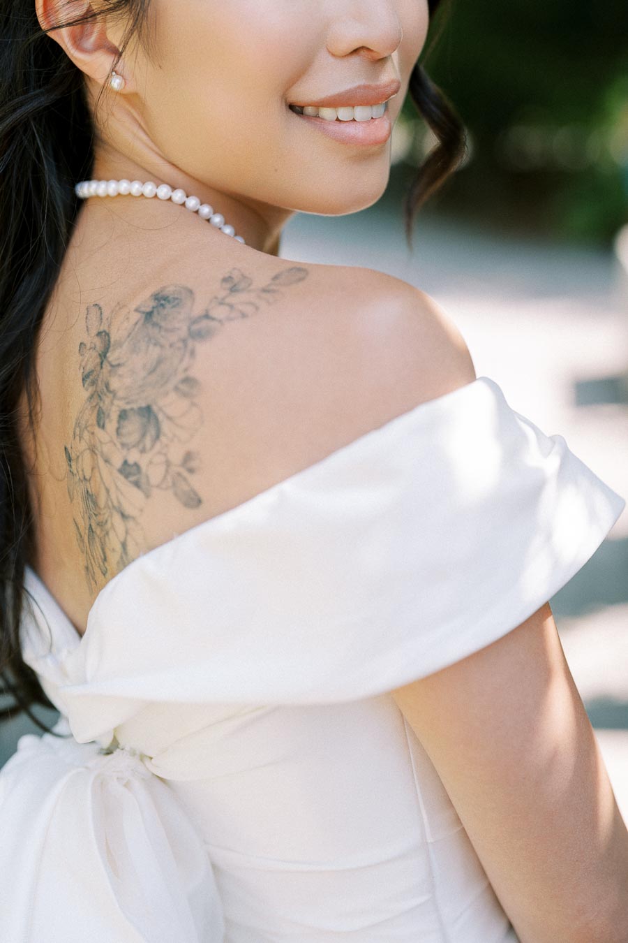 Bride with floral tattoo on shoulder wearing an off-the-shoulder white dress and pearl necklace, showcasing a beautiful bridal look.