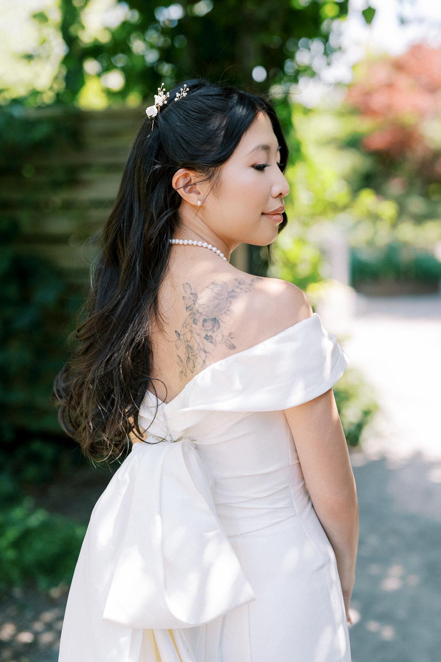 Young woman in an elegant white dress with floral shoulder tattoo, pearl necklace, and hair adorned with small flowers, standing in a garden setting.