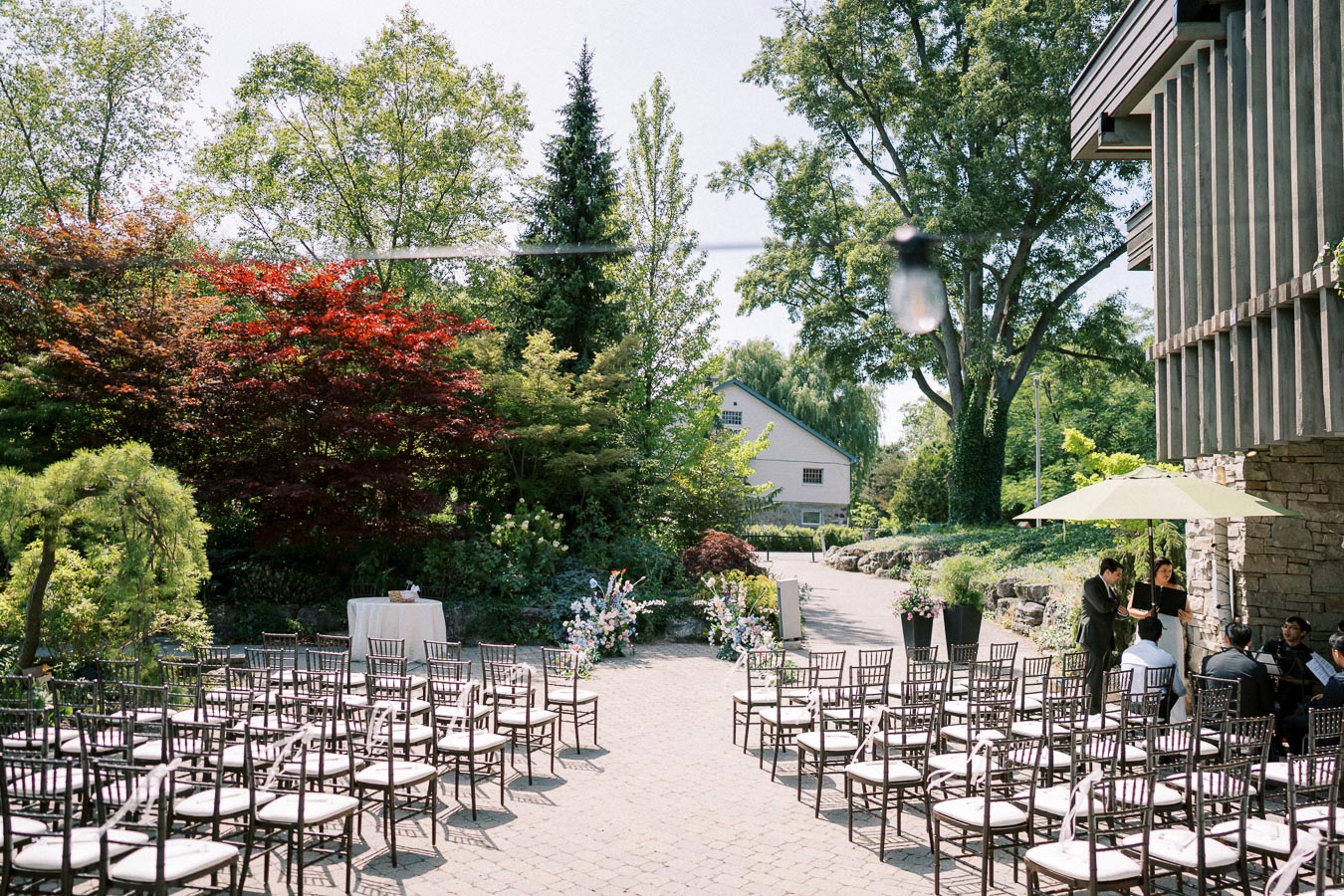 Outdoor wedding ceremony setup with rows of chairs and a table in a garden setting surrounded by lush trees and colorful foliage, with a building in the background.
