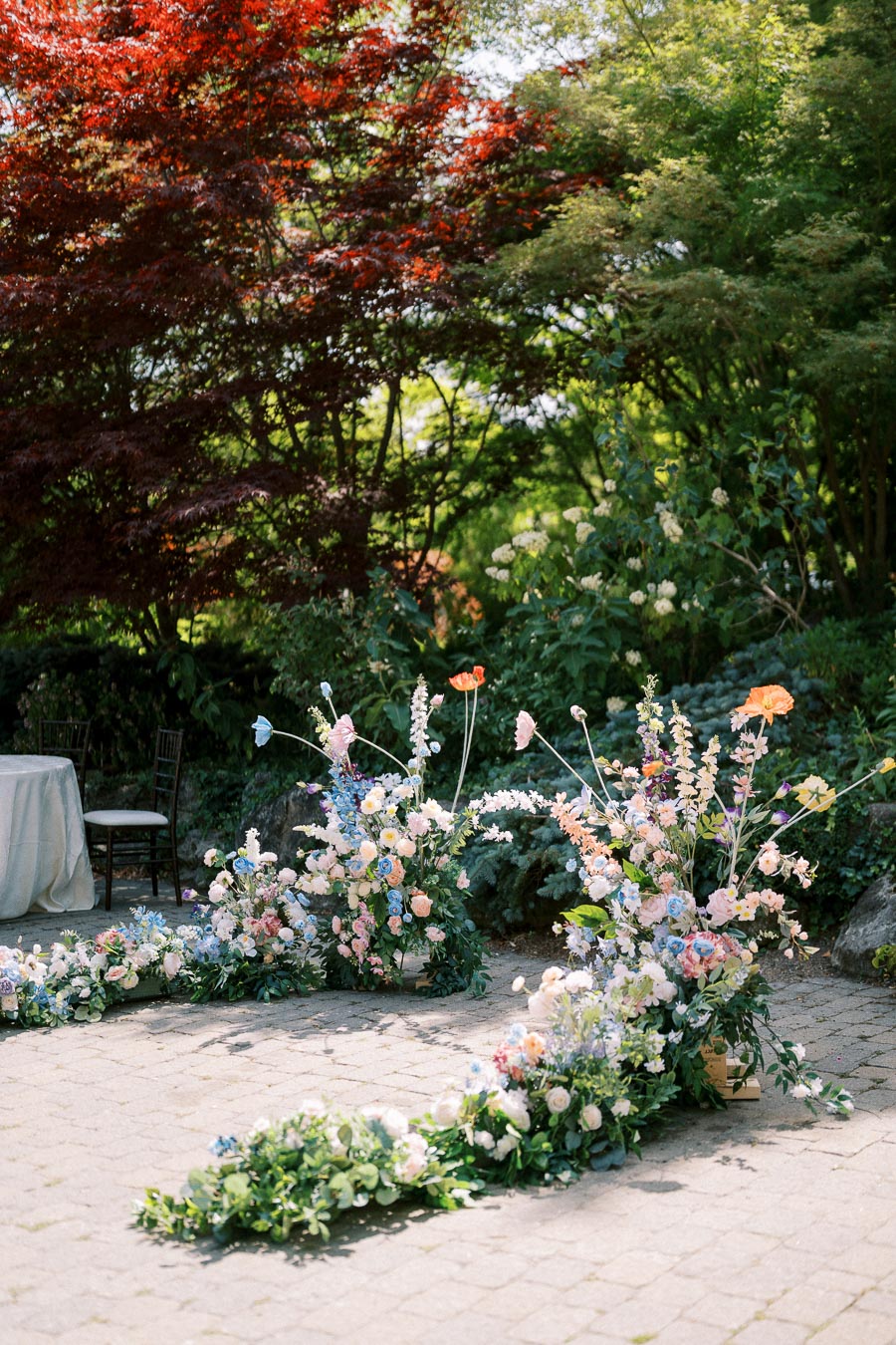 Elegant outdoor wedding setup with lush floral arrangements featuring pastel roses, delphiniums, and poppies. The charming garden setting is highlighted by vibrant green trees and dappled sunlight, creating a romantic and serene atmosphere.