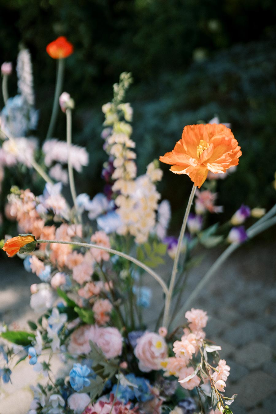 Colorful garden floral arrangement with vibrant orange poppies, surrounded by pastel-colored flowers and greenery, in a sunlit outdoor setting.