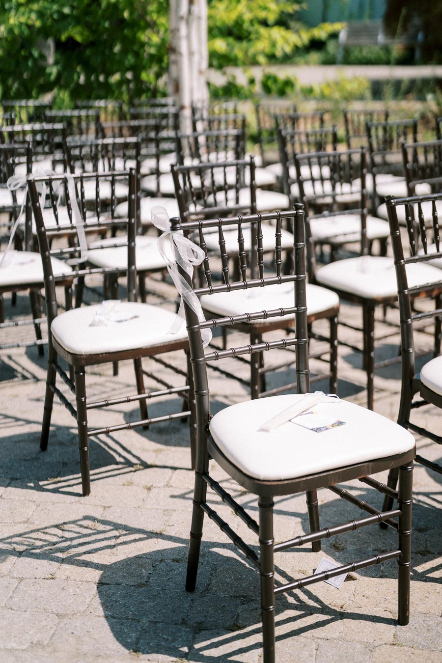 Rows of black Chiavari chairs with white cushions set up for an outdoor wedding ceremony on a sunny day, featuring greenery in the background.