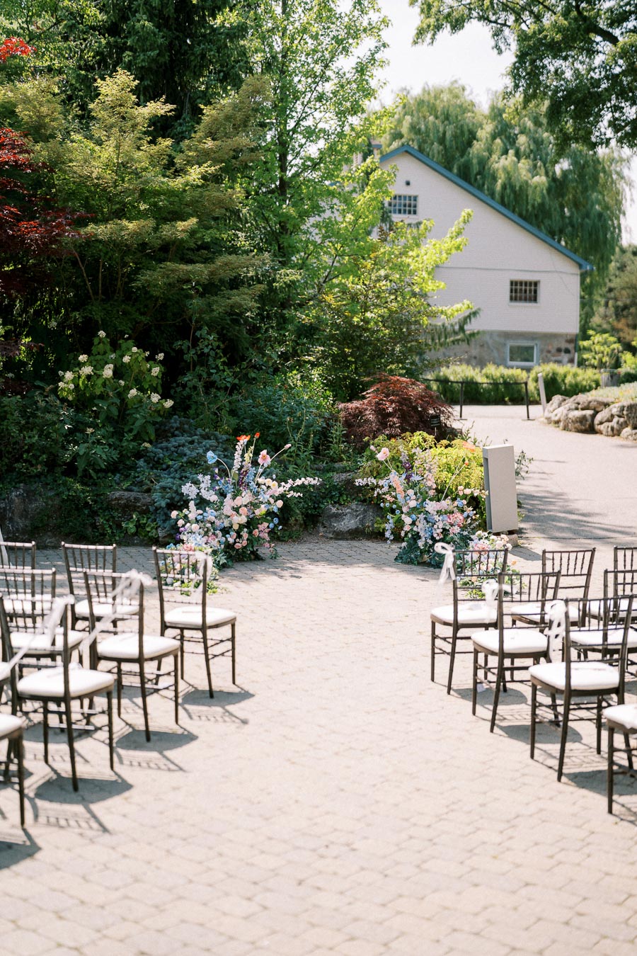 Outdoor wedding venue set up with elegantly arranged chairs and vibrant floral decorations, surrounded by lush greenery and a charming building in the background under a clear sky.