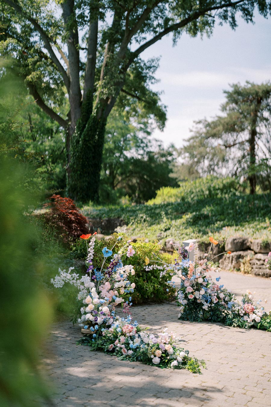 Outdoor garden setting with colorful flower arrangements and lush greenery under a large tree, perfect for a scenic wedding or special event backdrop.