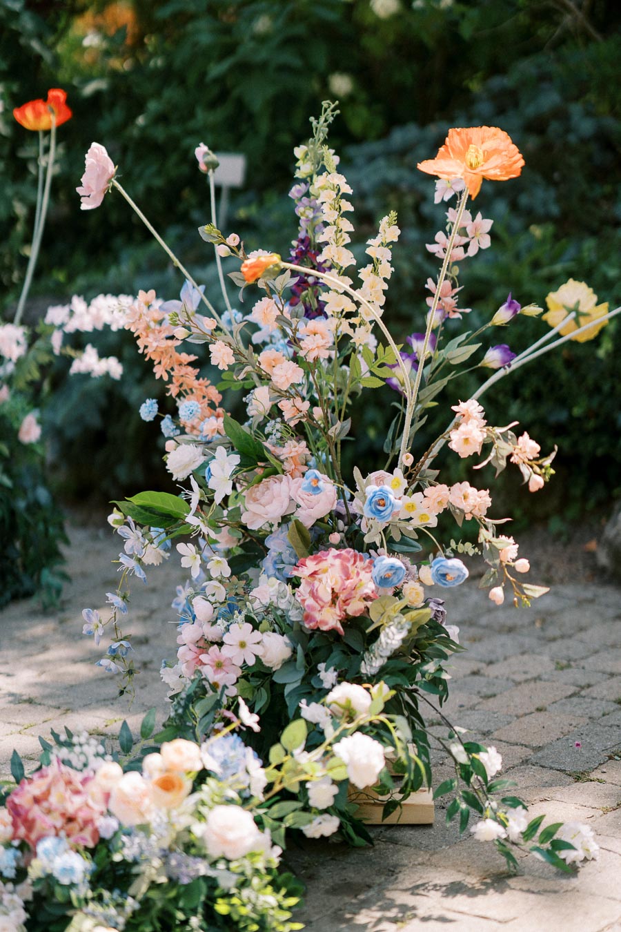 A vibrant arrangement of colorful wildflowers, including poppies and roses, elegantly displayed on a paved garden path, with green foliage and blurred greenery in the background.