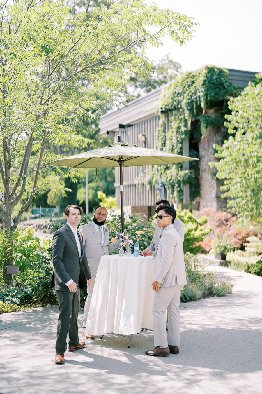 Group of men in suits at an outdoor event standing around a high table with a green umbrella, surrounded by lush greenery and a building covered in ivy.