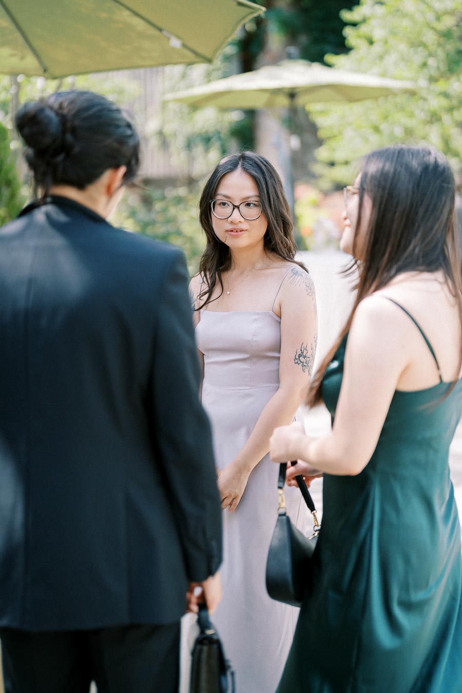Three people in formal attire engaged in conversation outdoors with green umbrellas and blurred foliage in the background.