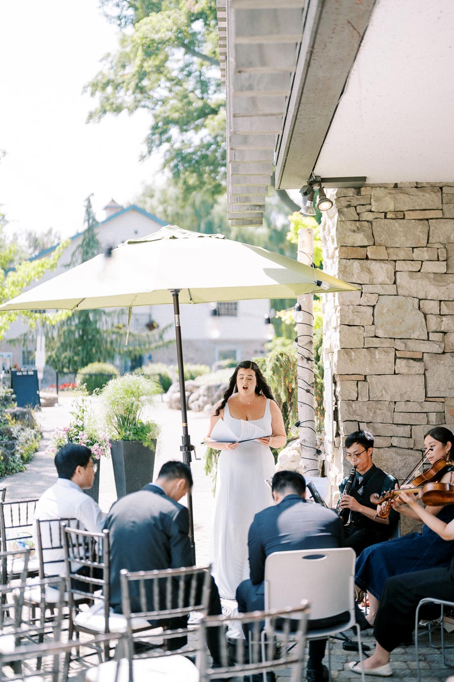 A woman in a white dress sings under a large umbrella during an outdoor event, accompanied by a seated small ensemble of musicians including a violinist and a clarinetist, against a backdrop of greenery and stone architecture.