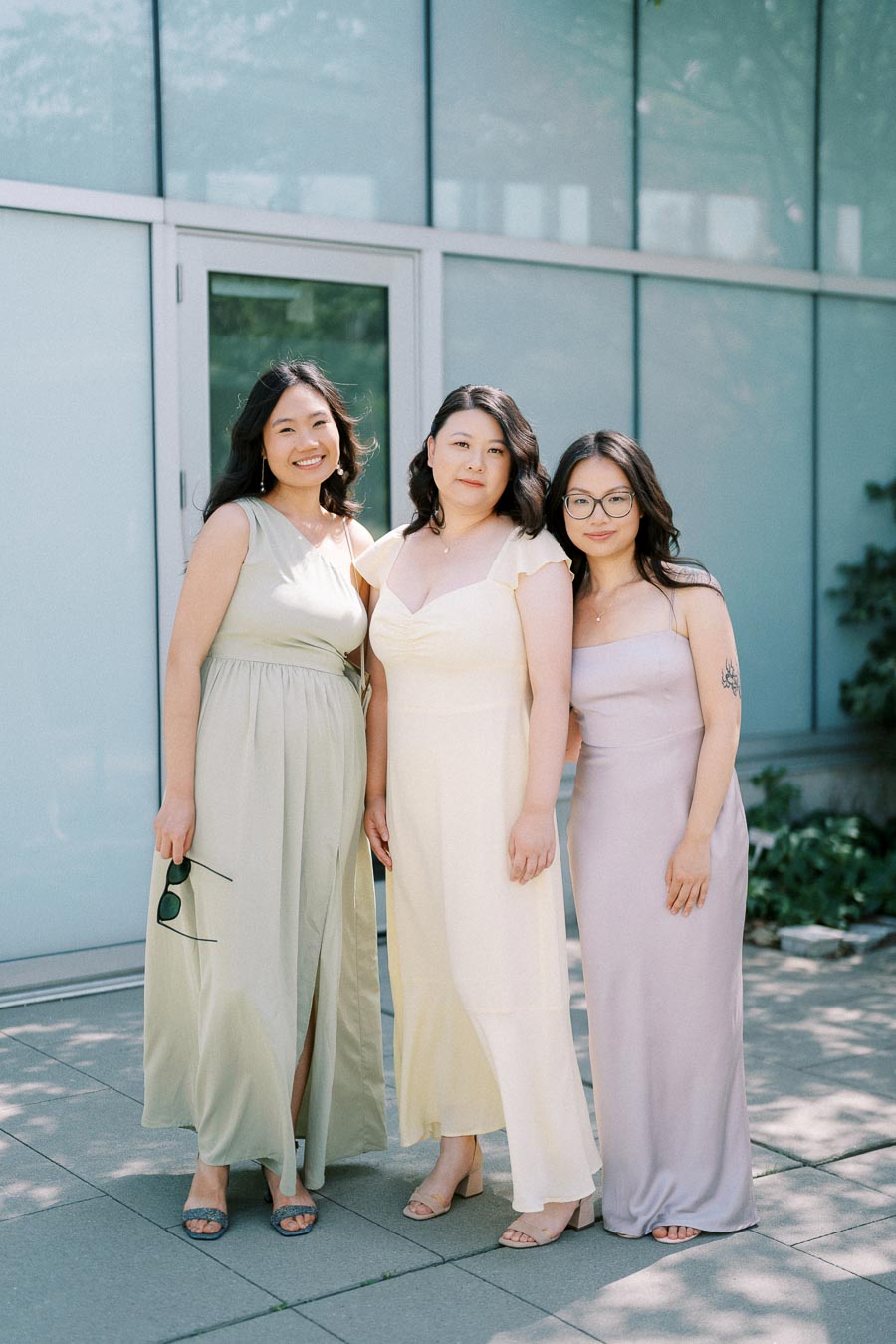 Three women in elegant pastel dresses stand outdoors in the sunlight, smiling with a glass building in the background.