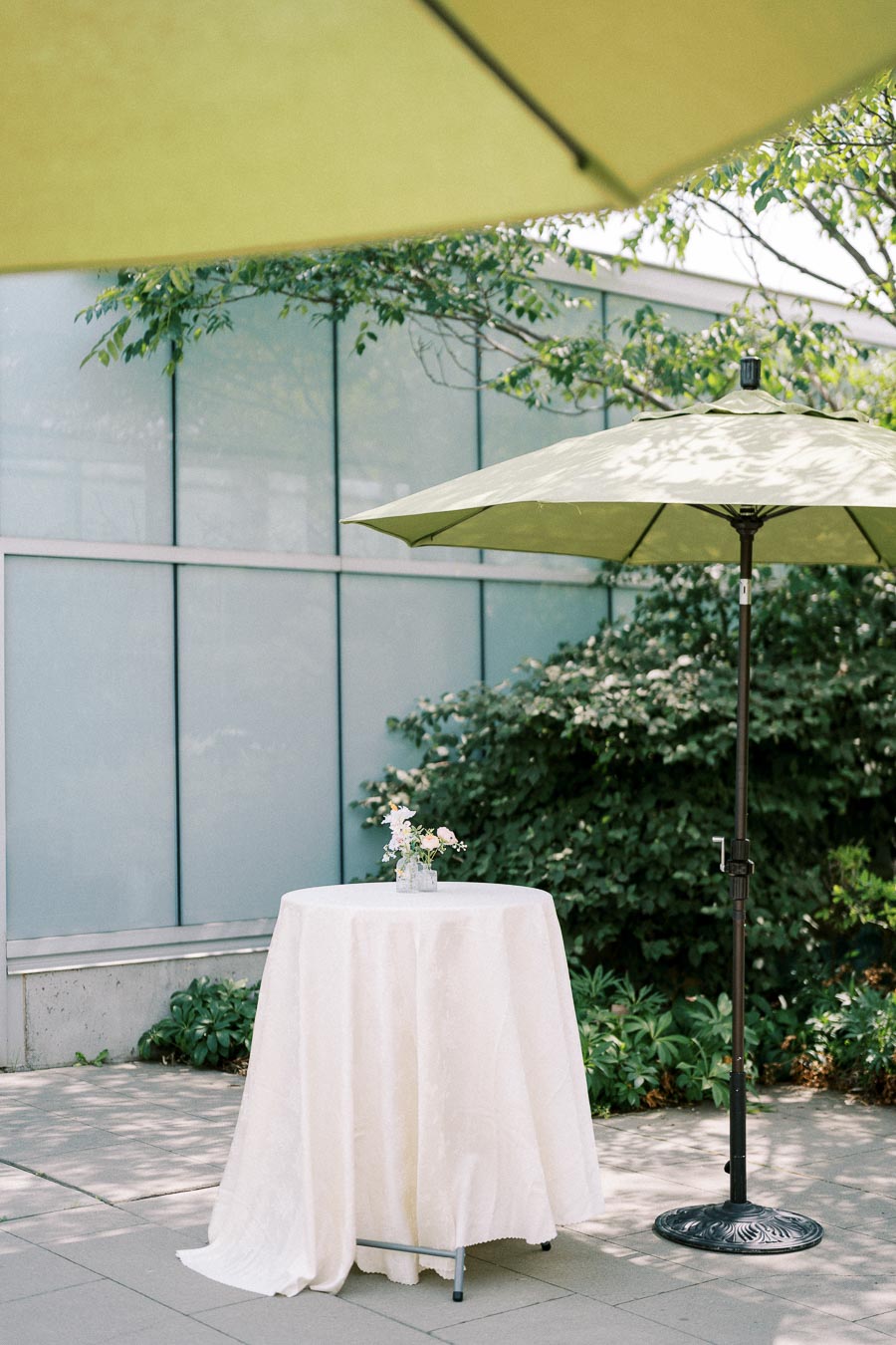Outdoor patio setting with a round table covered in a white tablecloth, adorned with a small floral arrangement. A green patio umbrella provides shade, with lush greenery in the background, creating a serene and inviting atmosphere.