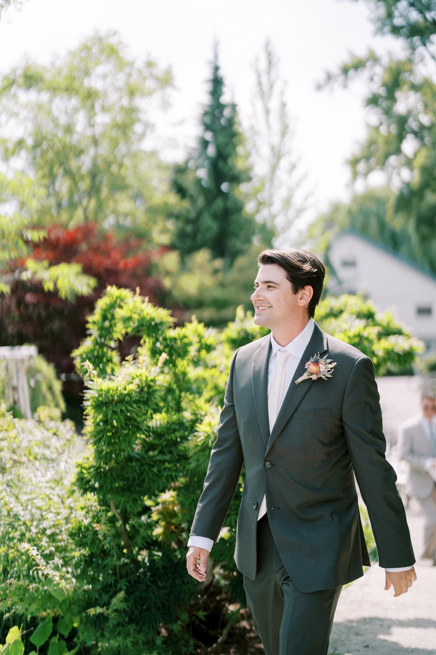 A smiling groom in a dark suit and white tie walks through a lush garden on a sunny day, with vibrant greenery and trees in the background.