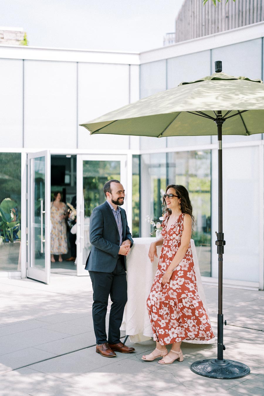 A man in a blue suit and a woman in a floral dress converse cheerfully at an outdoor event under a green umbrella. The backdrop features modern glass doors, and other attendees can be seen entering in the background.