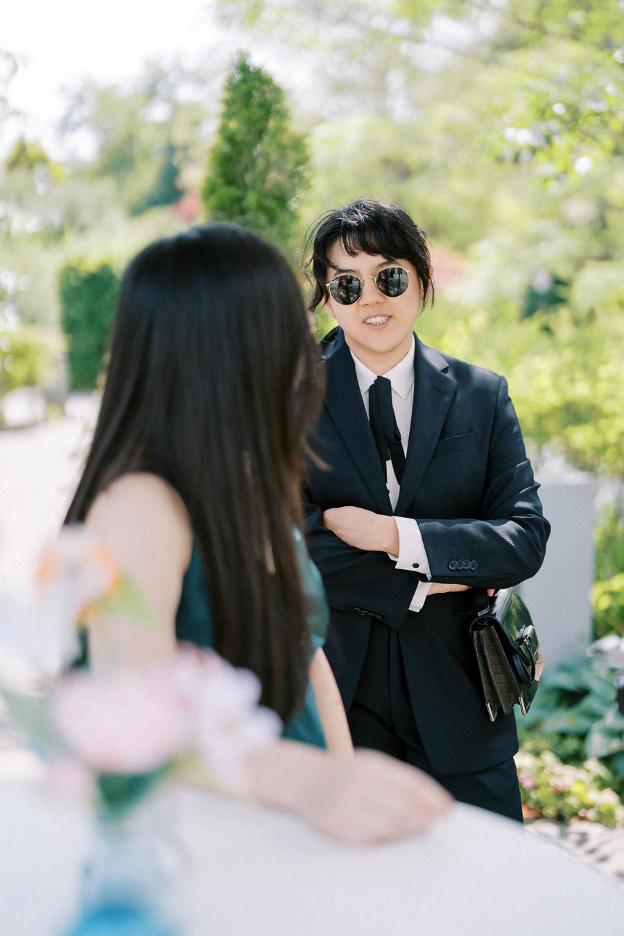 Two people having a conversation outdoors, one wearing a dark suit and sunglasses, with greenery in the background.