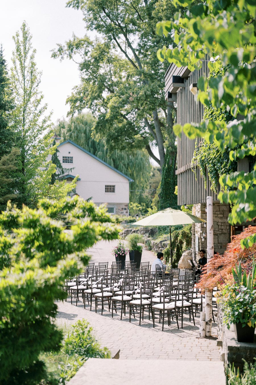 Outdoor garden venue with rows of chairs, lush greenery, stone building, and parasol on a sunny day.
