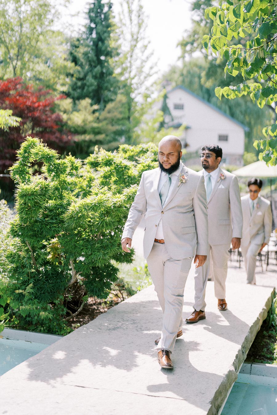 Groomsmen in light gray suits walking down a garden path at an outdoor wedding ceremony, with lush greenery and a rustic barn visible in the background.