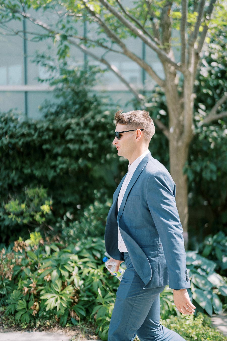 Man in a blue suit walking confidently outdoors in a sunny garden setting.