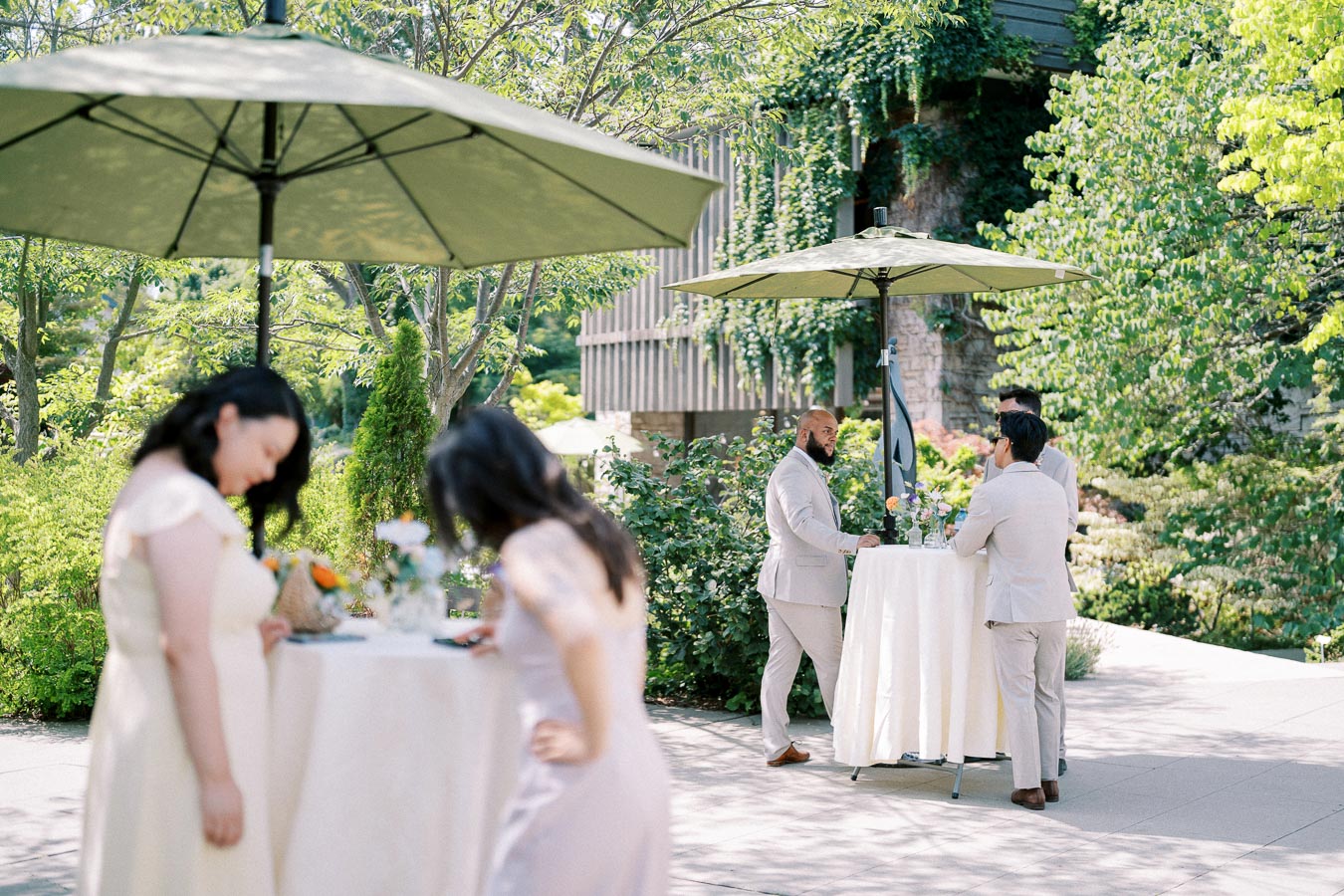 Outdoor garden gathering with guests in light attire standing under green umbrellas, surrounded by lush greenery and elegant white tablecloths.