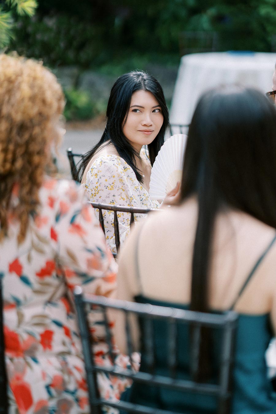 A woman in a floral dress holding a fan, sitting outdoors at a garden event, surrounded by other people in colorful attire.