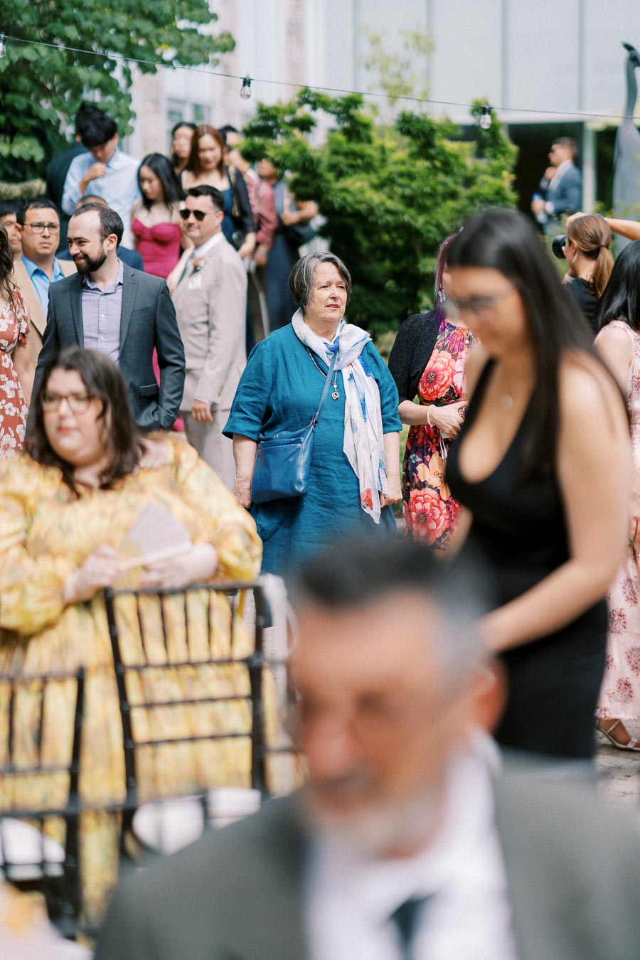 A group of people gathered at an outdoor event, with individuals dressed in formal and colorful attire, standing and sitting in front of lush greenery and a modern building backdrop.