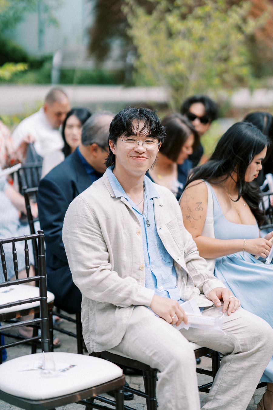 Smiling person seated outdoors at a formal event, wearing light-colored attire with other attendees in the background.