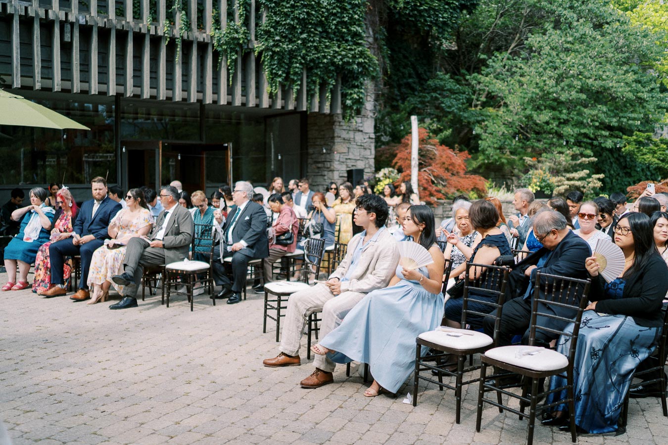 Outdoor wedding ceremony with guests seated on chairs in a garden setting, surrounded by greenery and a modern building facade.