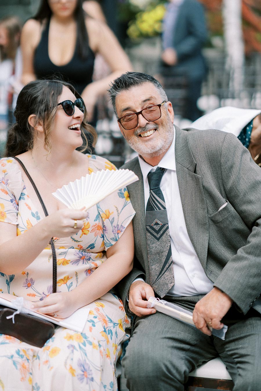 A woman in a floral dress and sunglasses smiles beside a man in a gray suit at an outdoor event.