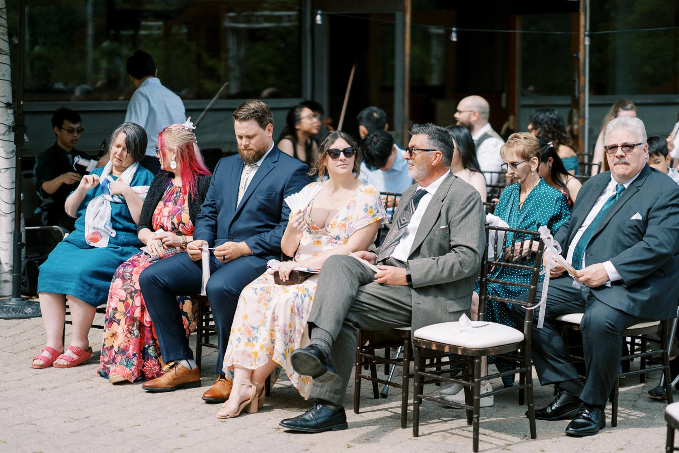 Outdoor wedding guests seated, dressed in formal attire, and interacting with each other.
