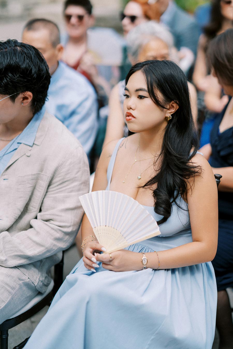 A woman in a light blue dress holding a hand fan, seated among a group of people at an outdoor event, with expressions of attentiveness.