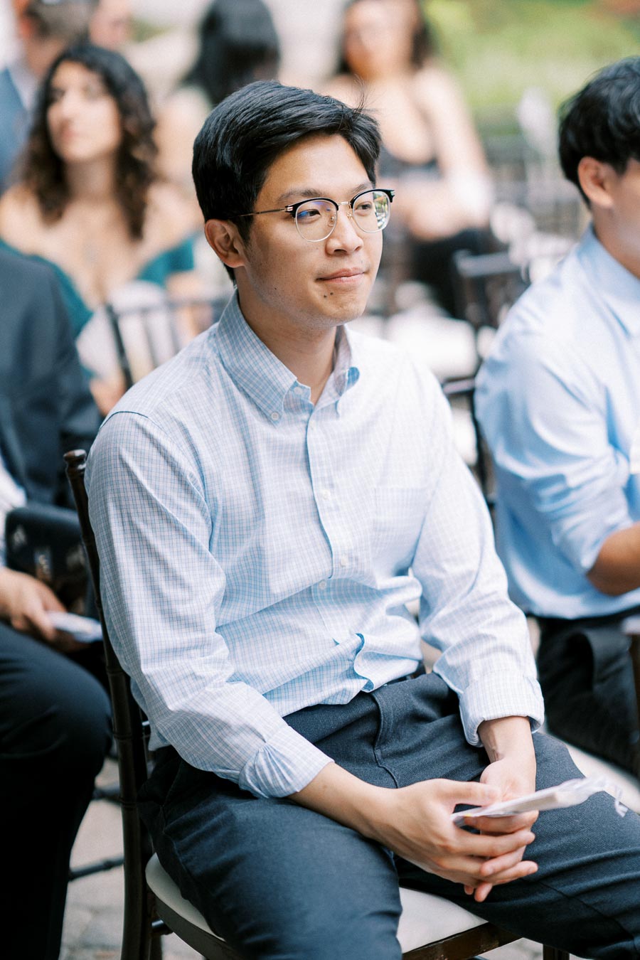 A person wearing glasses and a light checkered shirt seated attentively at an outdoor event, with other attendees visible in the blurred background.