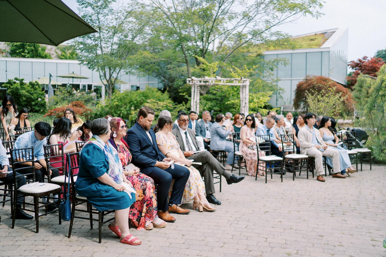 Outdoor wedding ceremony with guests seated on wooden chairs under a clear sky, surrounded by greenery and modern architecture in the background.