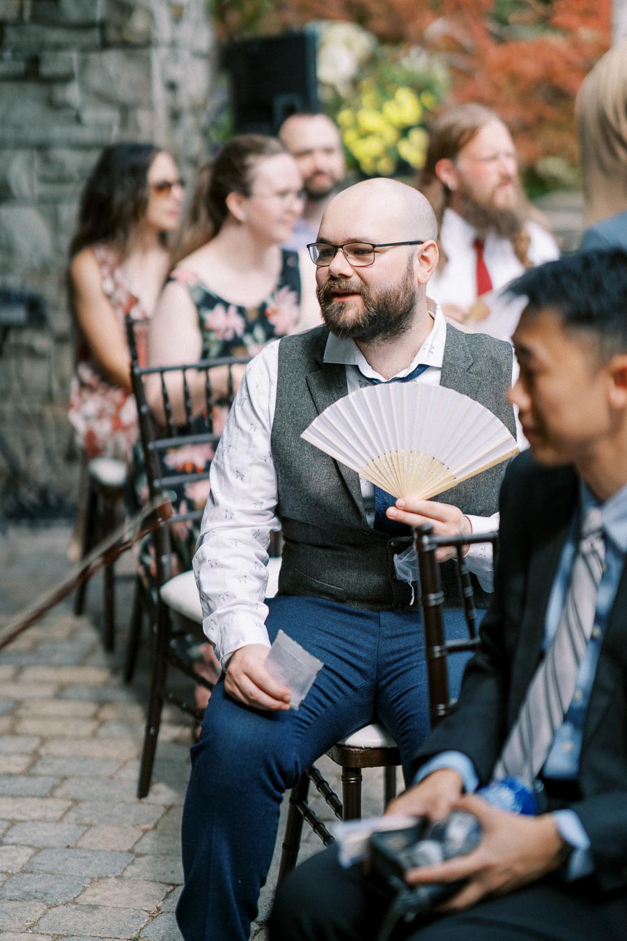 A well-dressed man with a beard and glasses holds a decorative hand fan while sitting among a group of people at an outdoor event.