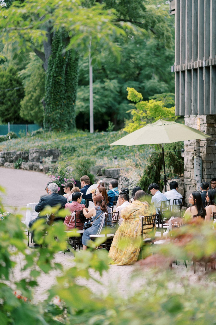 Outdoor garden wedding ceremony with seated guests under a green umbrella, surrounded by lush greenery and stone architecture.