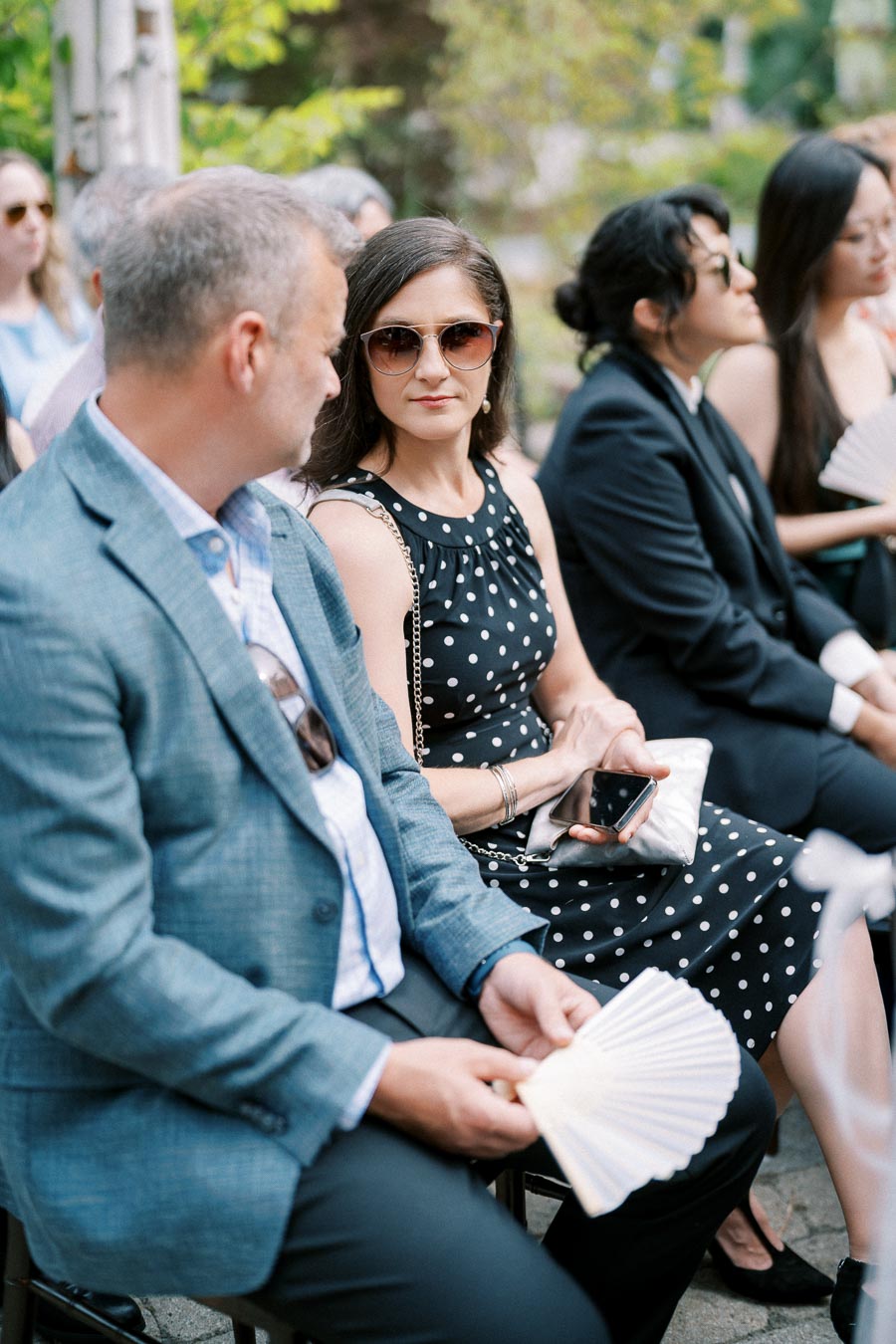 People sitting outdoors during a formal event, with a woman in a polka dot dress holding a phone and wearing sunglasses, surrounded by others in elegant attire.