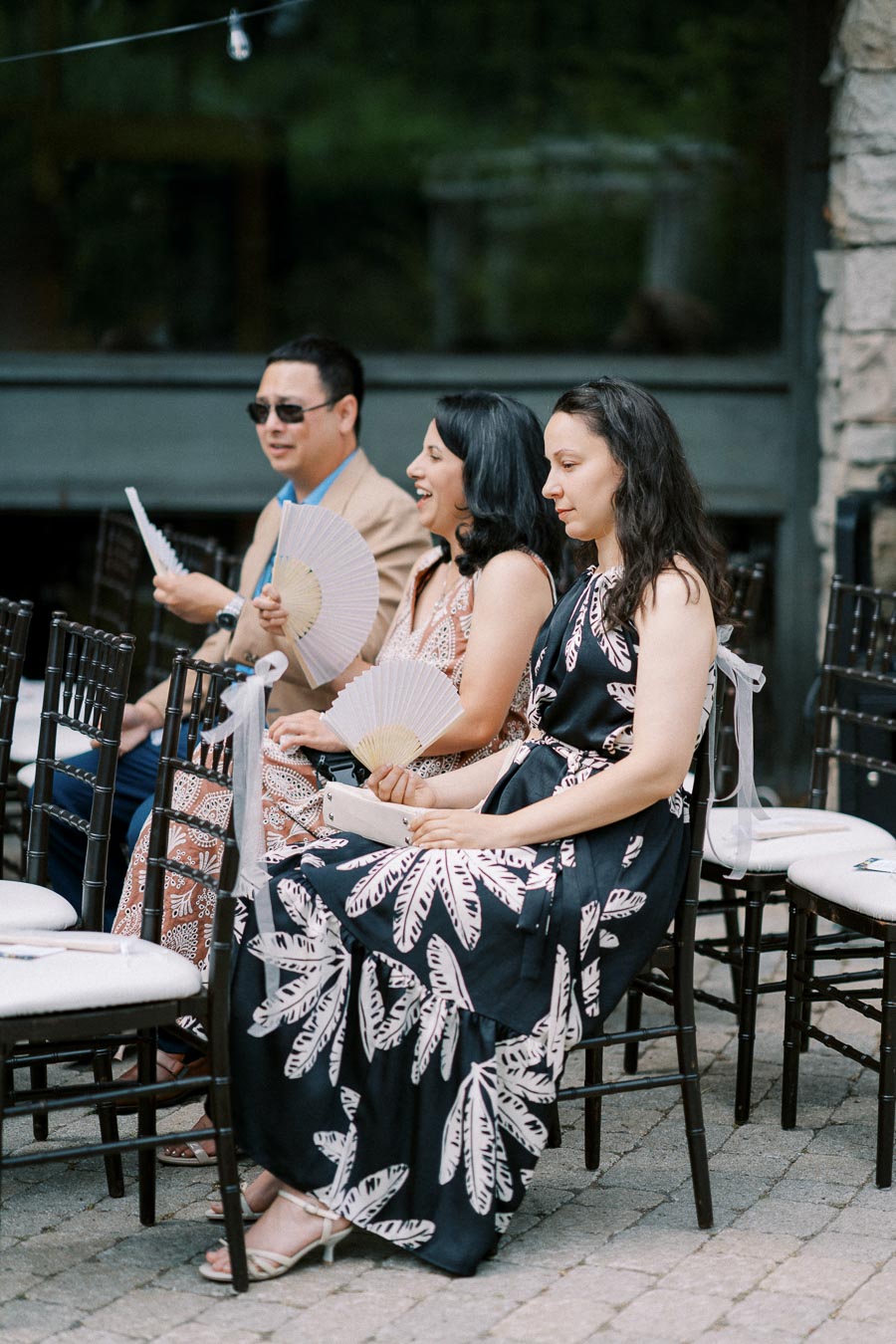 Guests seated at an outdoor wedding ceremony, holding paper fans, wearing elegant summer dresses and a suit, with black chairs arranged in rows.