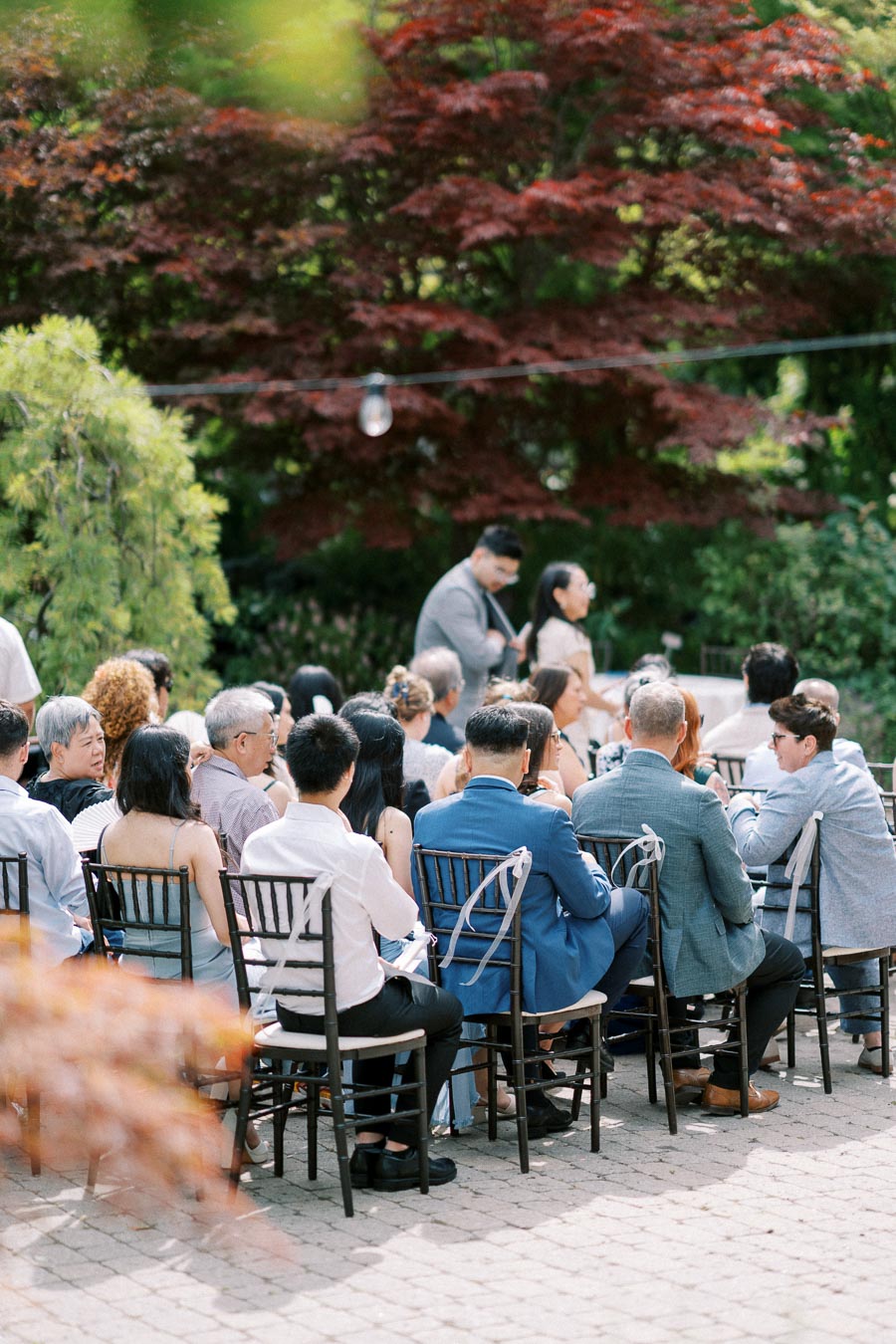 Outdoor wedding ceremony with guests seated on chairs, surrounded by lush greenery and red maple trees under soft lighting.