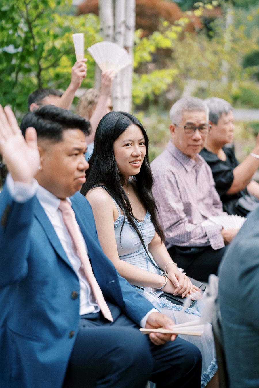 A group of people sitting outdoors at a formal event, with some holding fans. The setting is lush and green, suggesting a garden or outdoor ceremony. The people are dressed in formal attire, including a blue suit and a light blue dress.