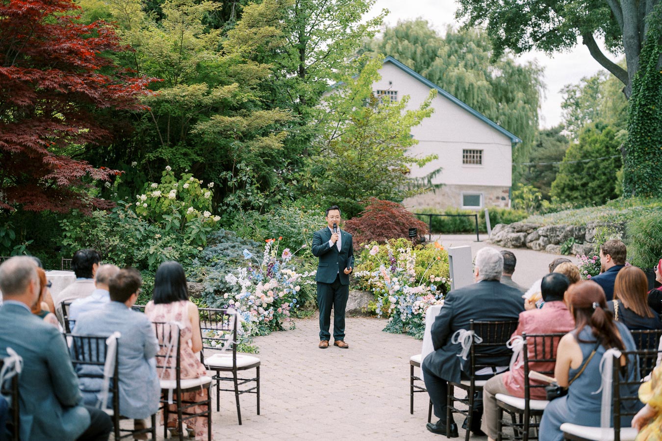 Outdoor wedding ceremony with guests seated in a garden setting, featuring a speaker in a suit standing in front of lush greenery and colorful floral arrangements, with a picturesque cottage in the background.