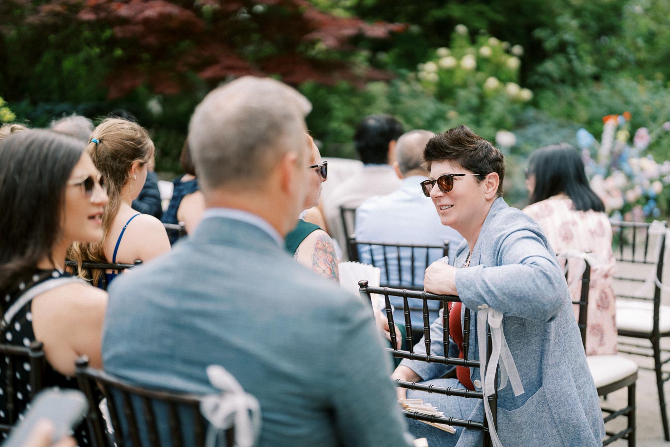Group of people seated outdoors at a garden event, wearing sunglasses and summer attire, engaging in conversation.