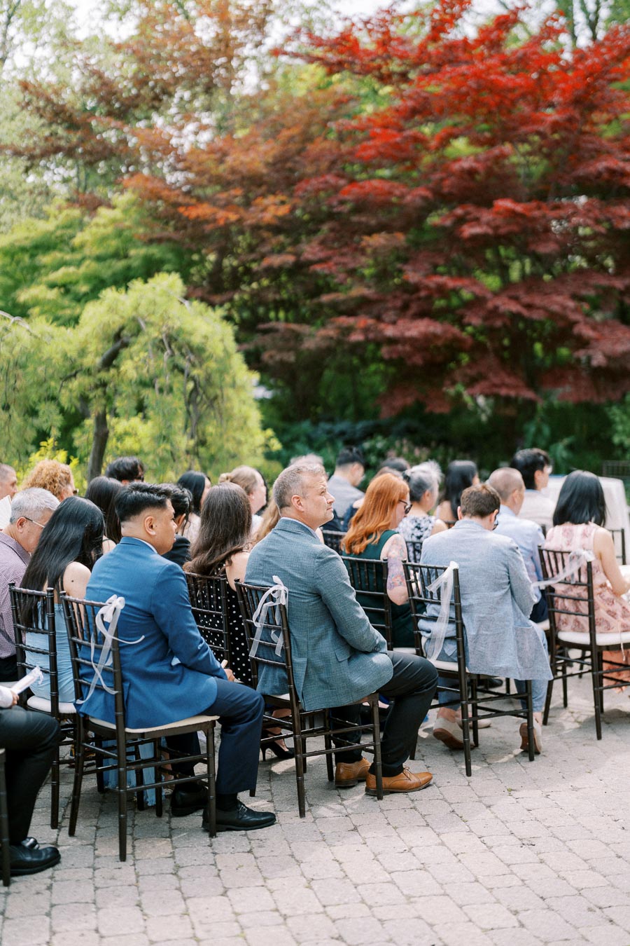 Guests seated outdoors at a wedding ceremony, surrounded by lush greenery and colorful foliage.