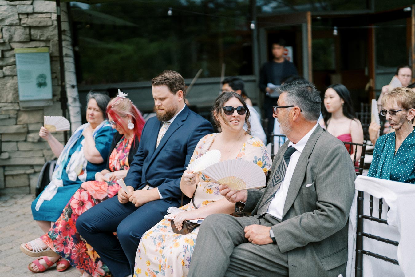 A group of people sits outdoors at a social event, dressed in colorful attire and suits, fanning themselves to stay cool.