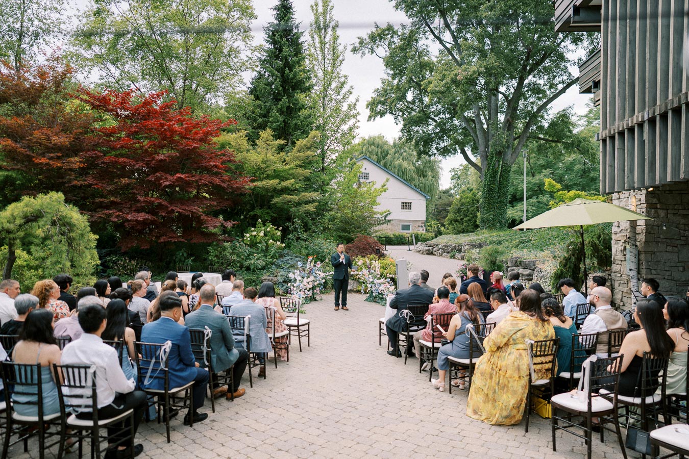 Outdoor wedding ceremony in a lush garden setting, featuring seated guests facing a speaker, surrounded by vibrant greenery and floral arrangements, with a stone pathway and rustic building in the background.
