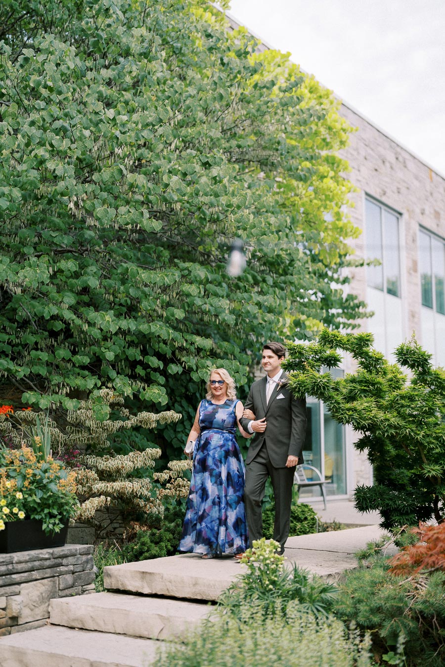 Elderly woman in a blue floral dress and young man in a suit walking arm-in-arm down a stone path surrounded by lush greenery and vibrant flowers, with a modern building in the background.