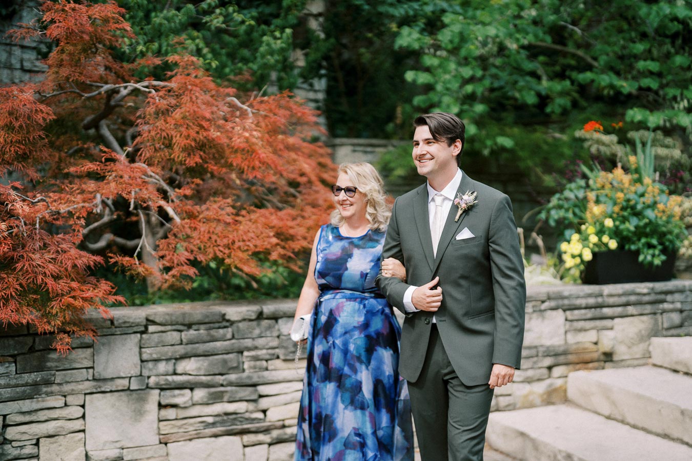 A smiling couple walks arm-in-arm in an outdoor garden setting, with a background of lush greenery and vibrant orange foliage. The man wears a sharp grey suit with a boutonnière, and the woman sports a blue patterned dress and sunglasses.