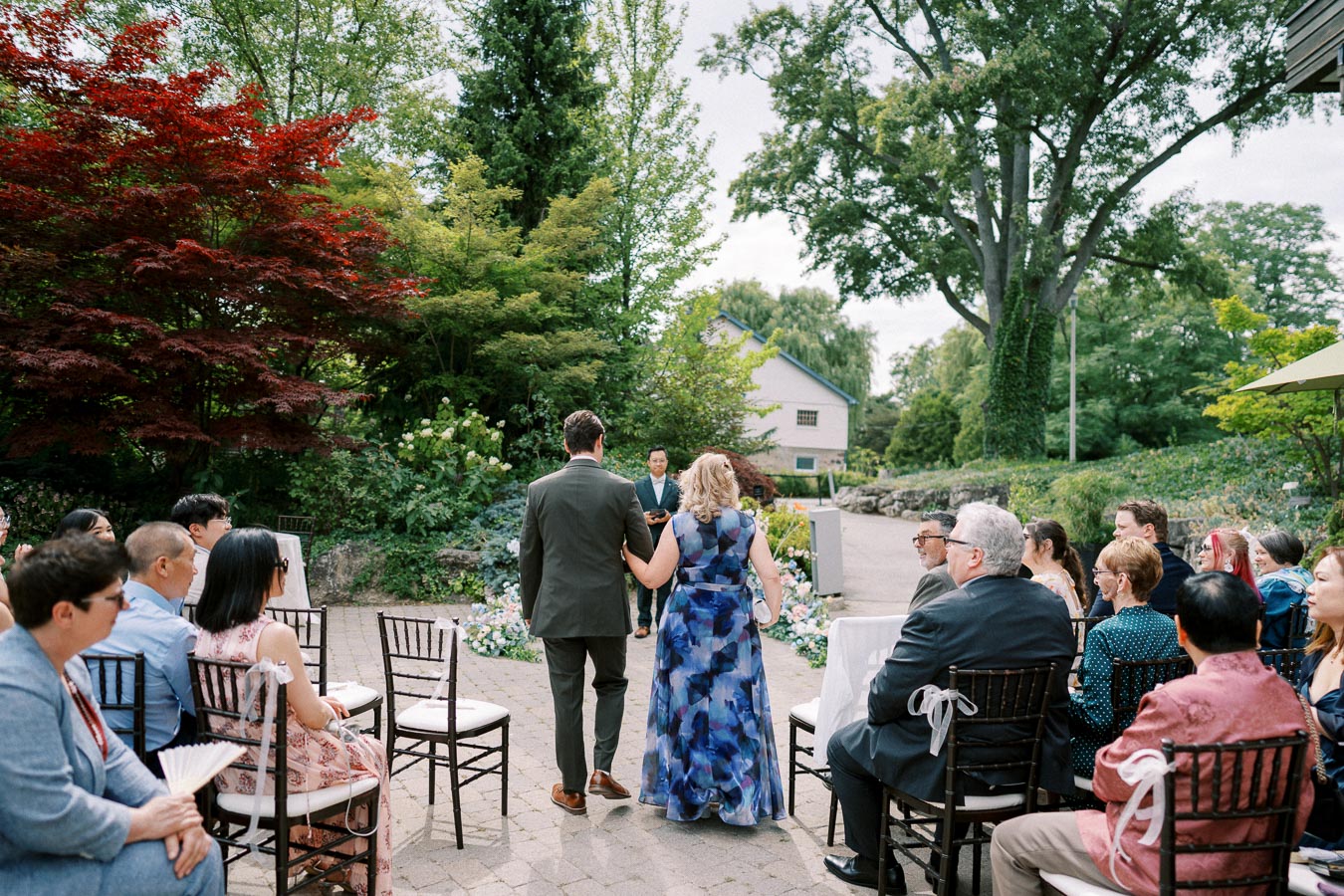 Outdoor garden wedding ceremony with a bride in a blue floral dress and a groom in a suit walking down the aisle, surrounded by seated guests, lush greenery, and vibrant flowers.